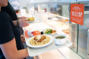 Person hält gefülltes Tablett in Kantine und schaut auf eine Plakette von United Against Waste